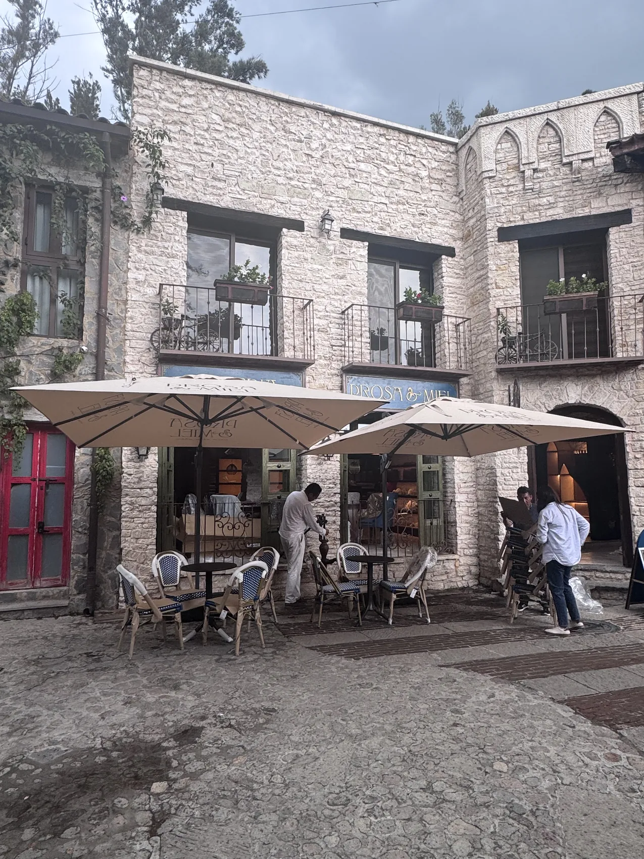 Terraza exterior de la librería con sombrillas de lona, mesas de hierro y la fachada de piedra del pueblo al fondo.
